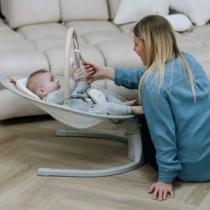 Babyschaukel Elite in Grau mit Spielbogen und zwei Spielzeugen.